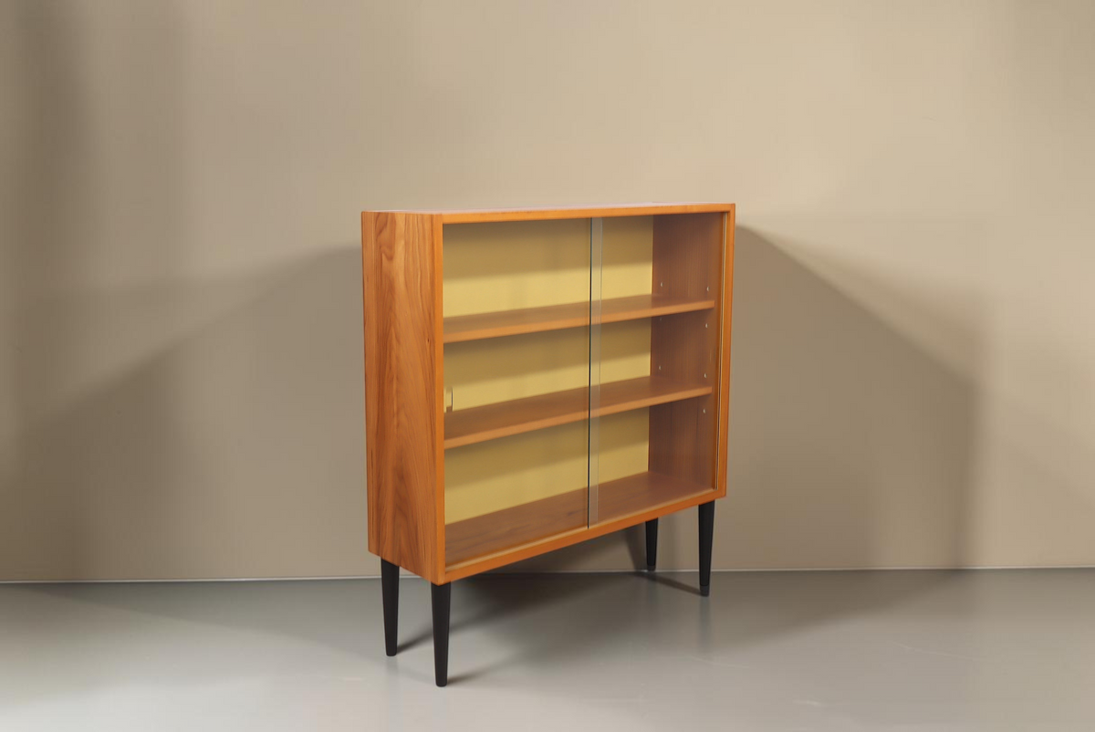 Vintage Space Hellerau Mid Century Vitrine aus Holz mit Glasschiebetüren und senfgelber Rückwand auf schwarzen Beinen